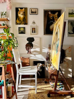 Artist studio with easel, paintings of cats on walls, white chair, and houseplants in sunlight
