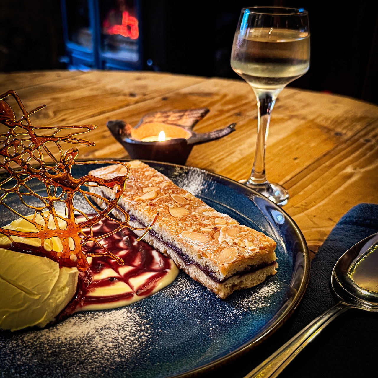 Slice of Bakewell tart on a plate with powdered sugar, sauce, and a glass of white wine