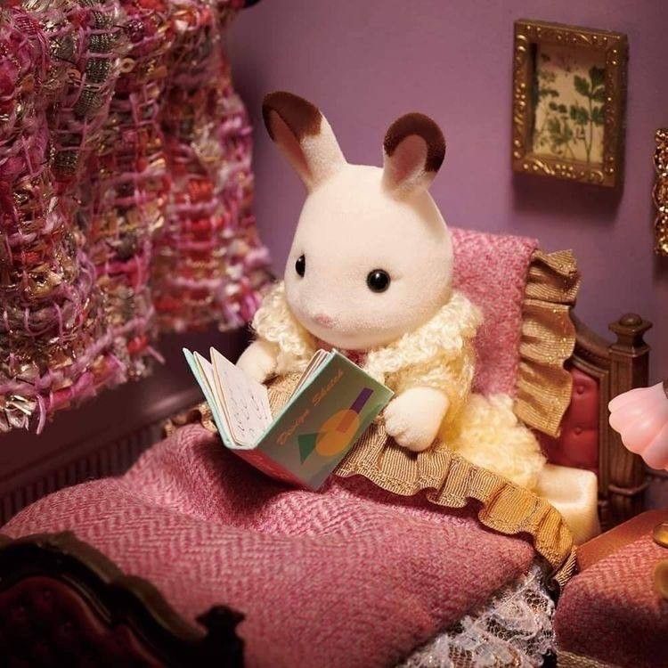Rabbit doll reading a book while sitting on a pink bed in a miniature bedroom