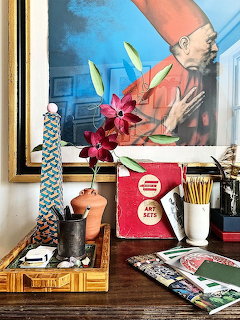 Art supplies, flowers, and books on a wooden desk in front of a framed portrait painting