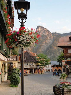 Street view of alpine village with hanging flower baskets, traditional buildings, and mountain peak in background