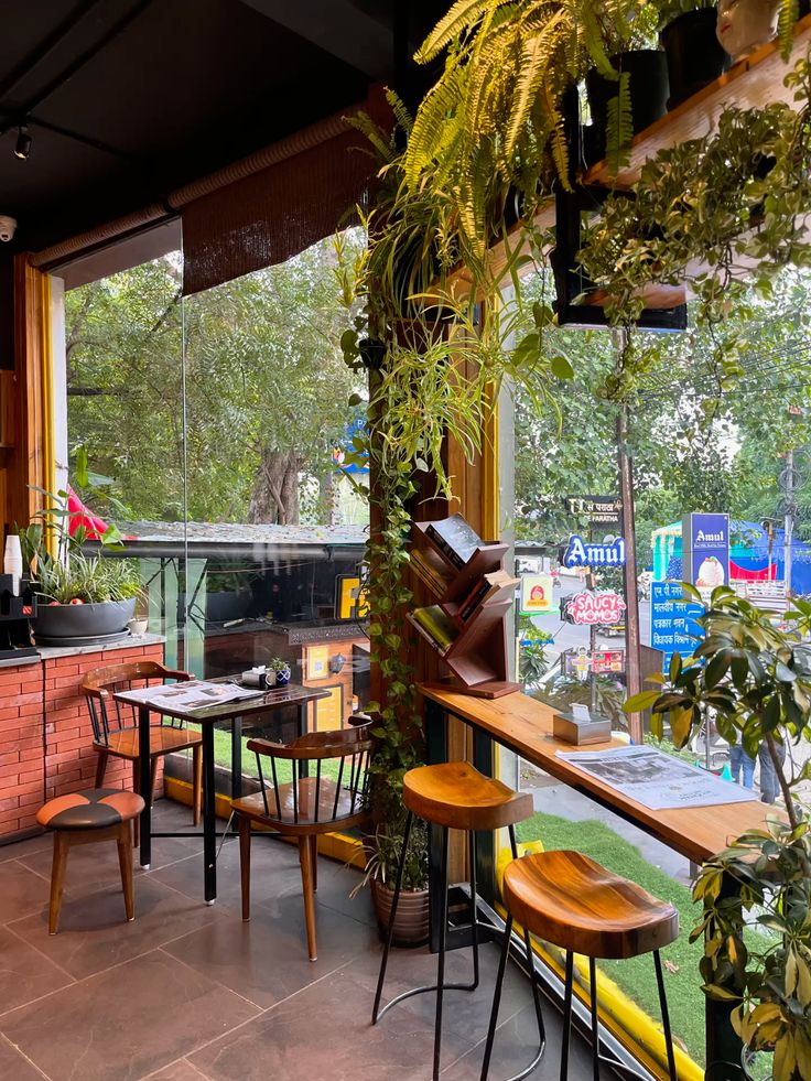 Cozy cafe terrace with abundant green plants, wooden bar stools, and open windows overlooking trees
