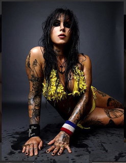 Woman with dark hair and arm tattoos sitting on dark floor in black top and yellow shorts