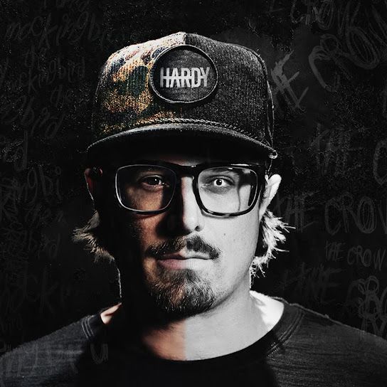 Man wearing glasses and a HARDY cap in a dark studio portrait