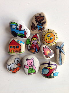 Nine smooth stones painted with colorful cartoon characters and objects including a train, sun, wolf, pig, and helicopter.