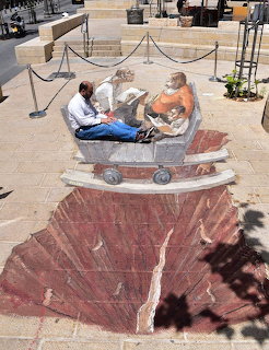 Two people posing with 3D chalk art creating illusion of sitting on edge of painted canyon on street pavement