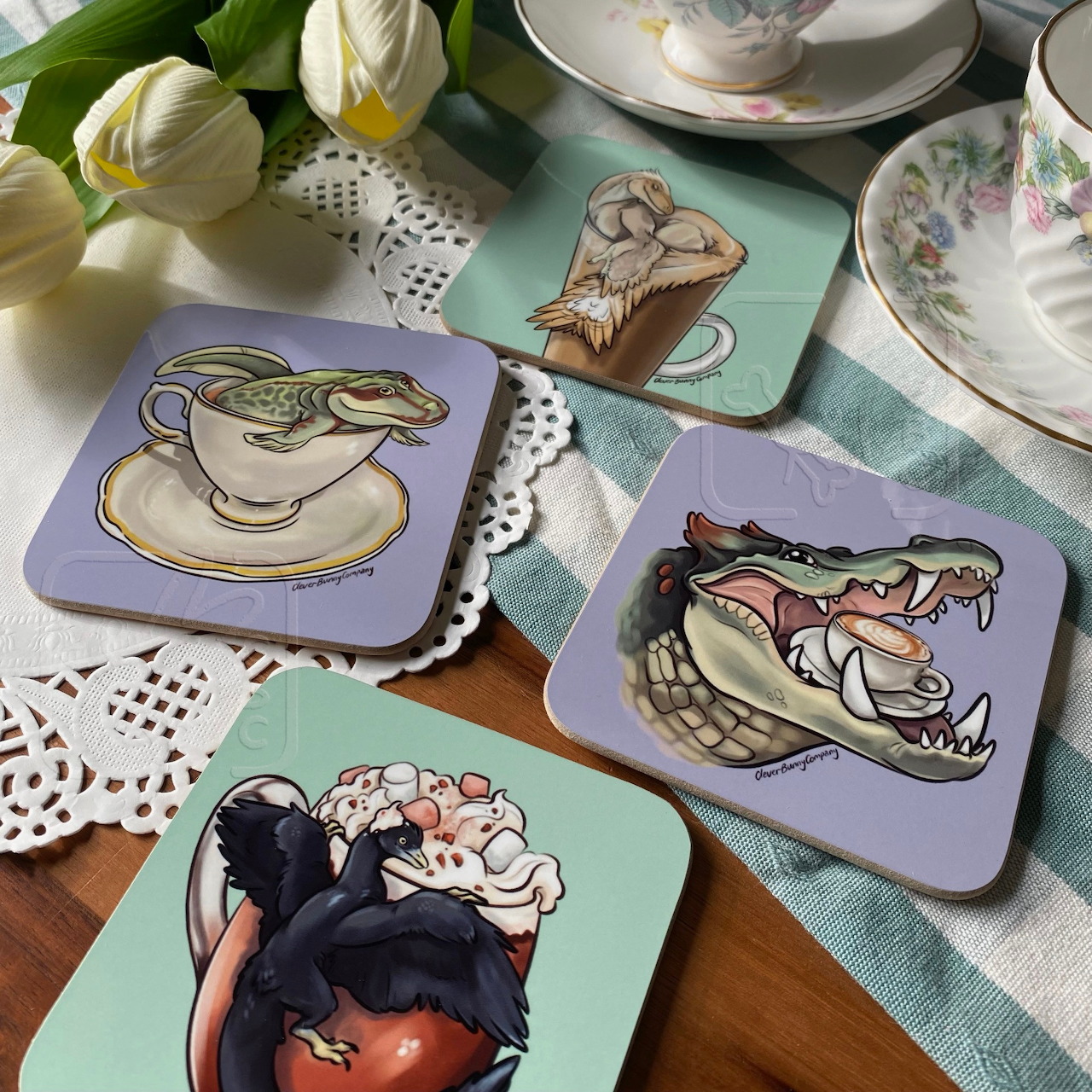 Five illustrated coasters showing dinosaurs in teacups arranged on table with white china and flowers