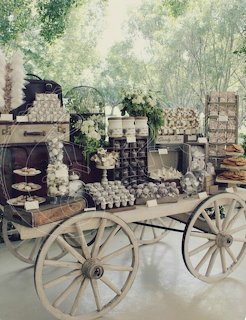 Vintage white wooden cart with tiered dessert display, cupcakes, flowers, and rustic suitcases outdoors among trees