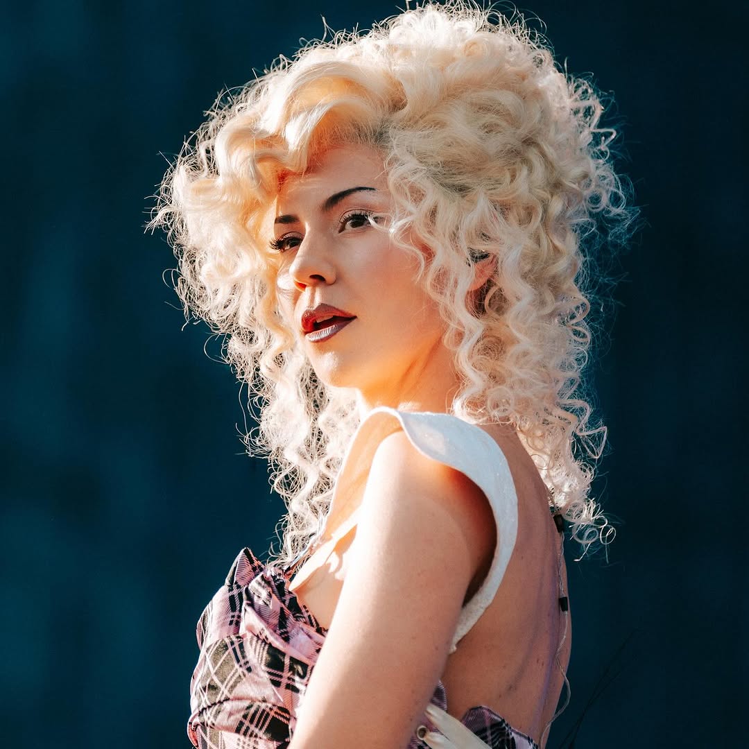 Side profile of woman with voluminous platinum blonde curly hair against teal background