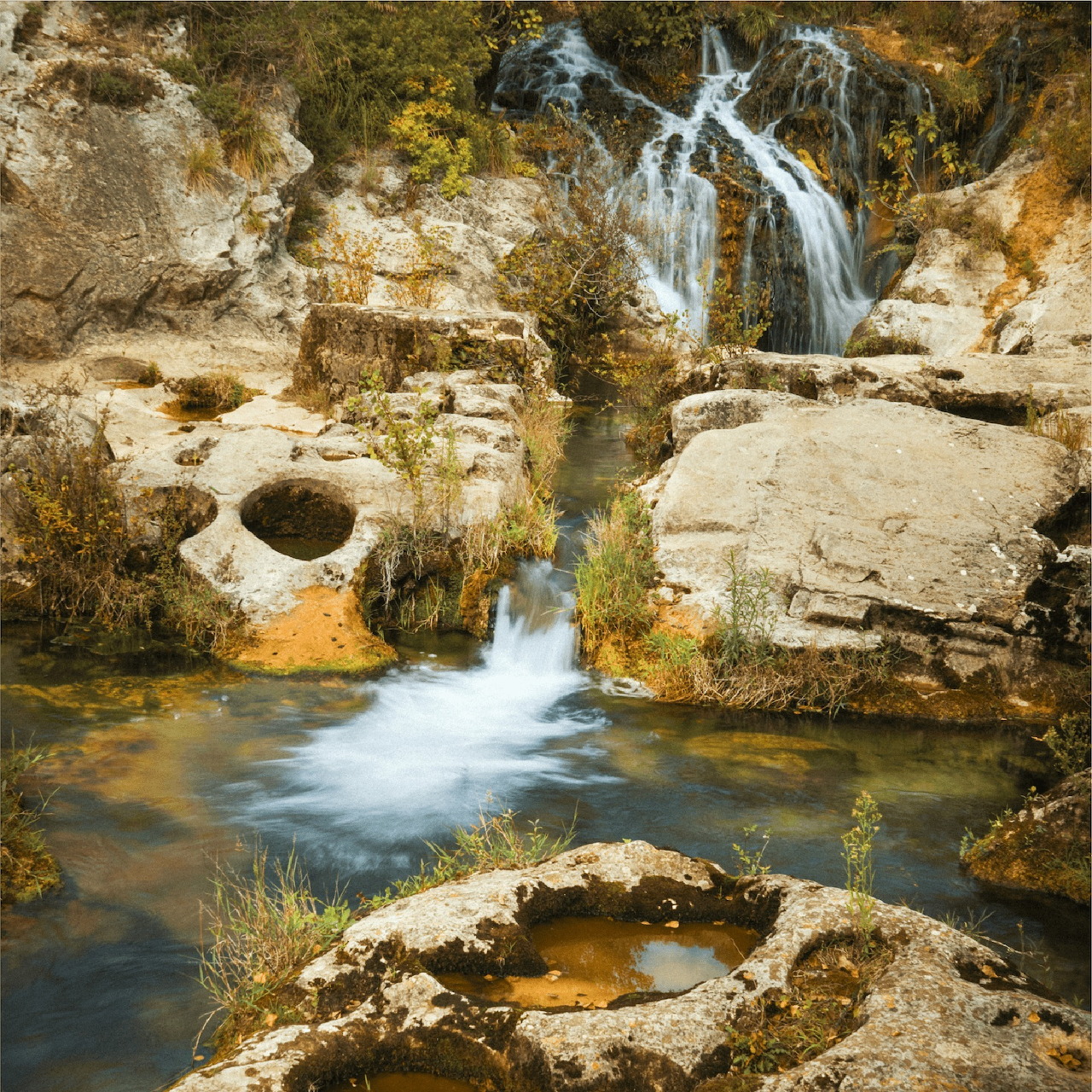 Small waterfall flowing into a rocky pool with smooth stone formations and dry grasses