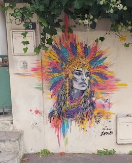 Street art portrait of woman with colorful radiating headdress and paint drips on building wall with plants