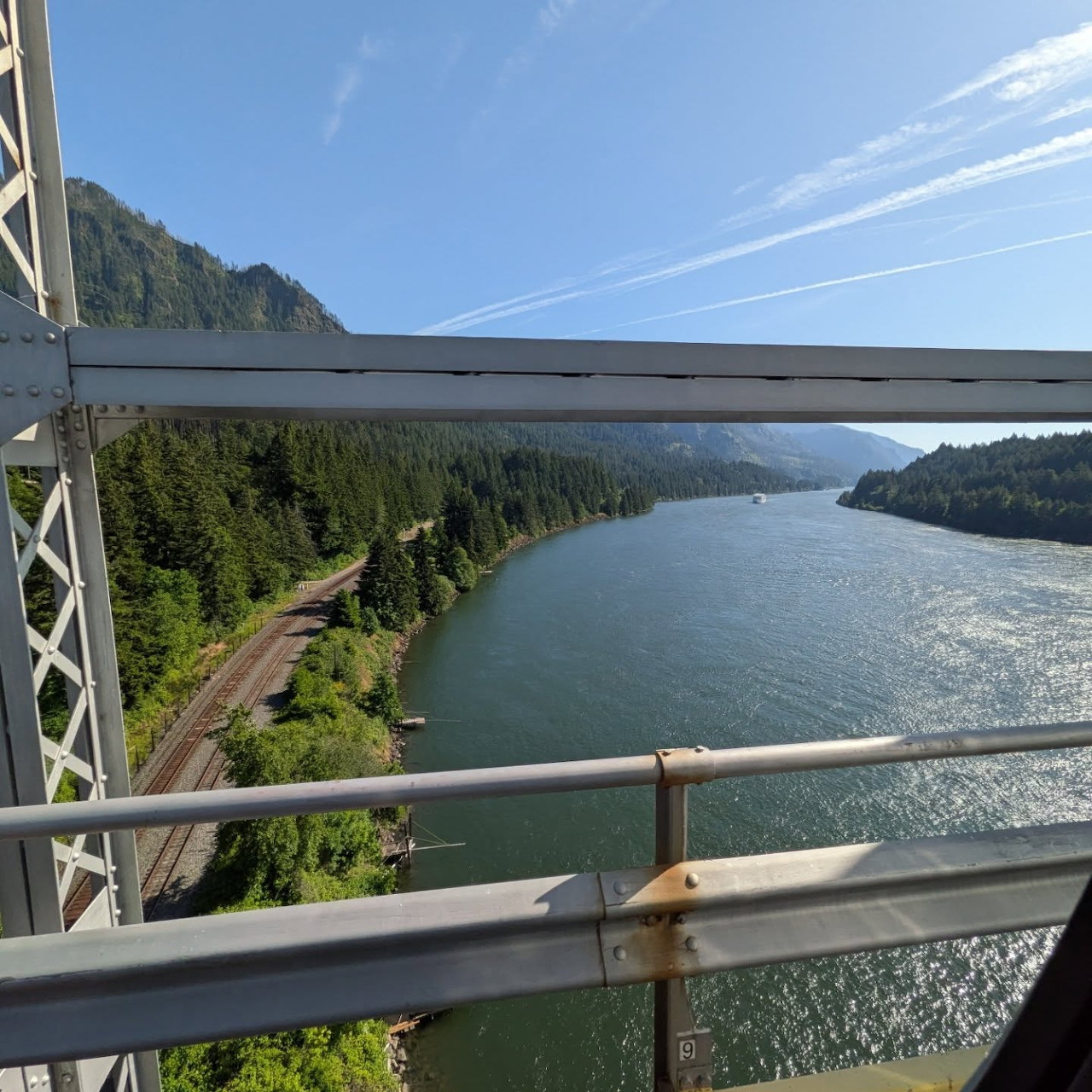 View from a steel bridge over a wide river with forested mountains and railroad tracks along the shore