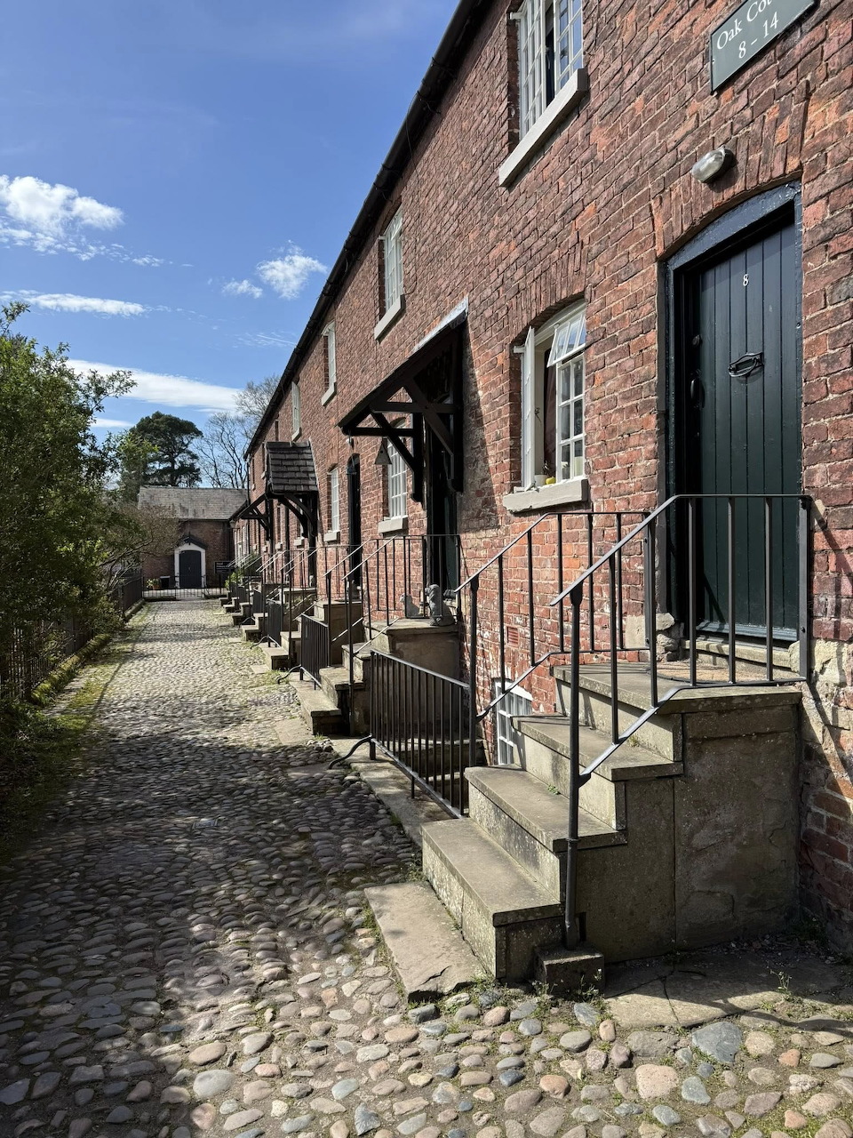 Row of red brick cottages with cobblestone path, dark green doors, and metal railings on a sunny day