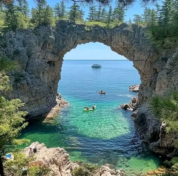 Stone arch formation over turquoise water with kayakers and pine trees