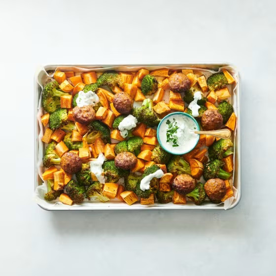 Sheet pan with roasted sweet potato, broccoli, meatballs, and white sauce drizzle from above