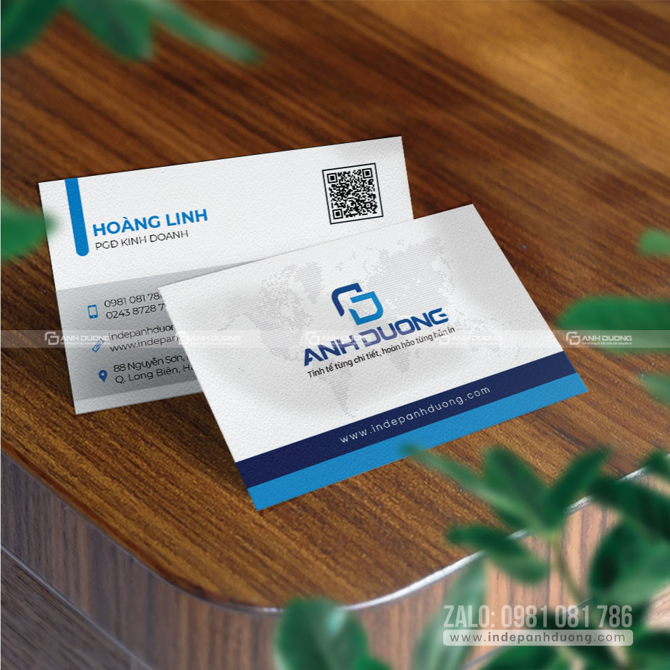 Two business cards with blue design on a wooden table, framed by blurred green leaves