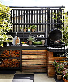 Outdoor kitchen with wooden counters, stored firewood, round BBQ grill, and potted plants under dark pergola