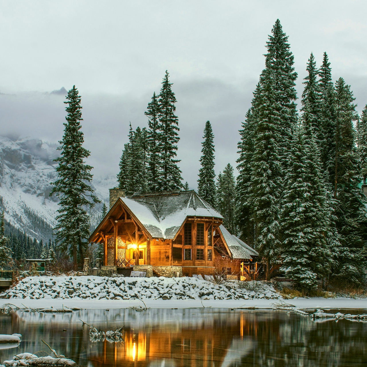 Warmly lit wooden cabin by a snowy lake with tall pine trees and misty mountains