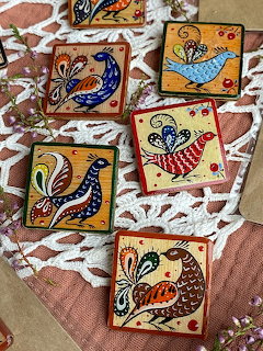 Five square ornaments featuring stylized birds in blue, red, and yellow with decorative patterns on lace