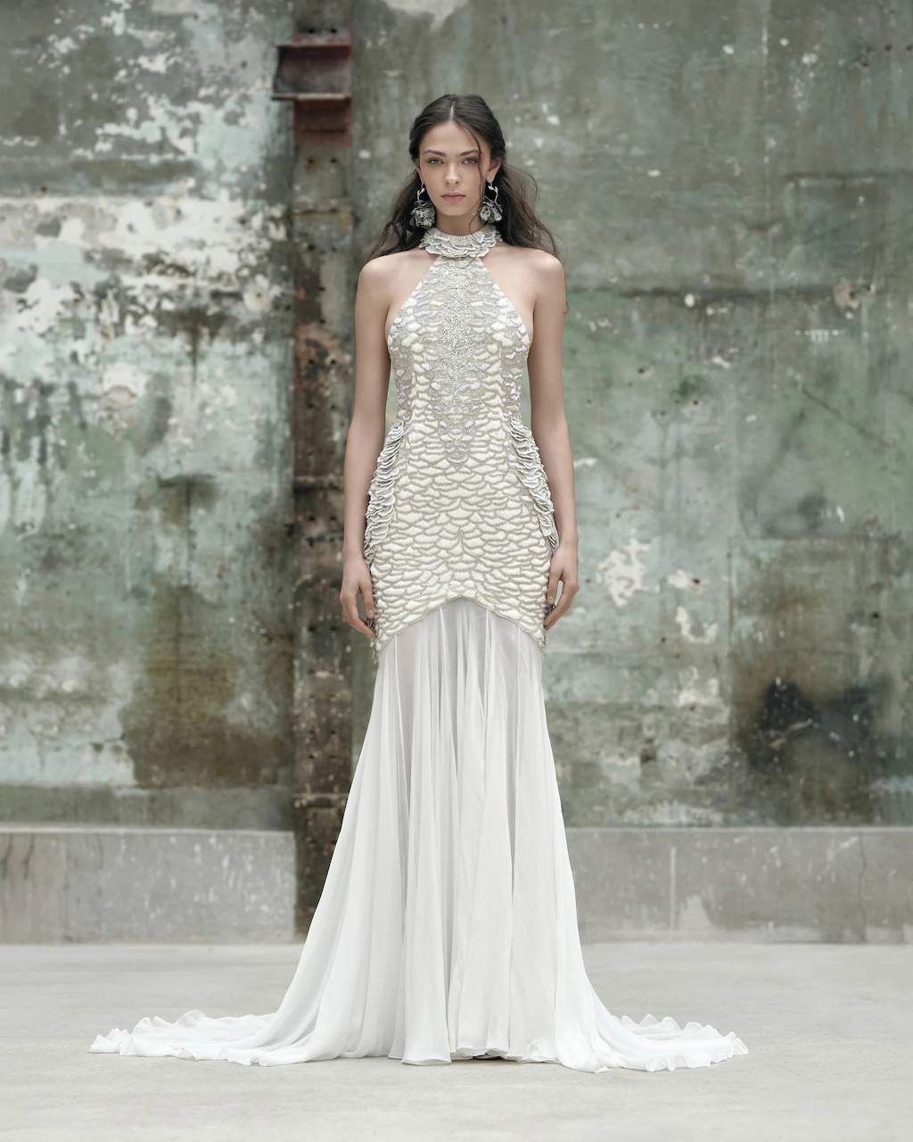 Model wearing white beaded halter bridal gown with flowing skirt against distressed wall