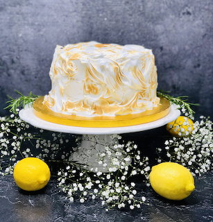 Lemon meringue cake on a stand with two lemons and small white flowers on a dark surface