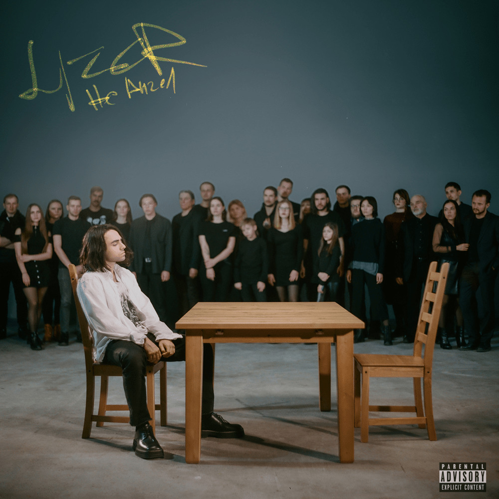 Man sitting at wooden table with empty chair, crowd standing in background, album cover style