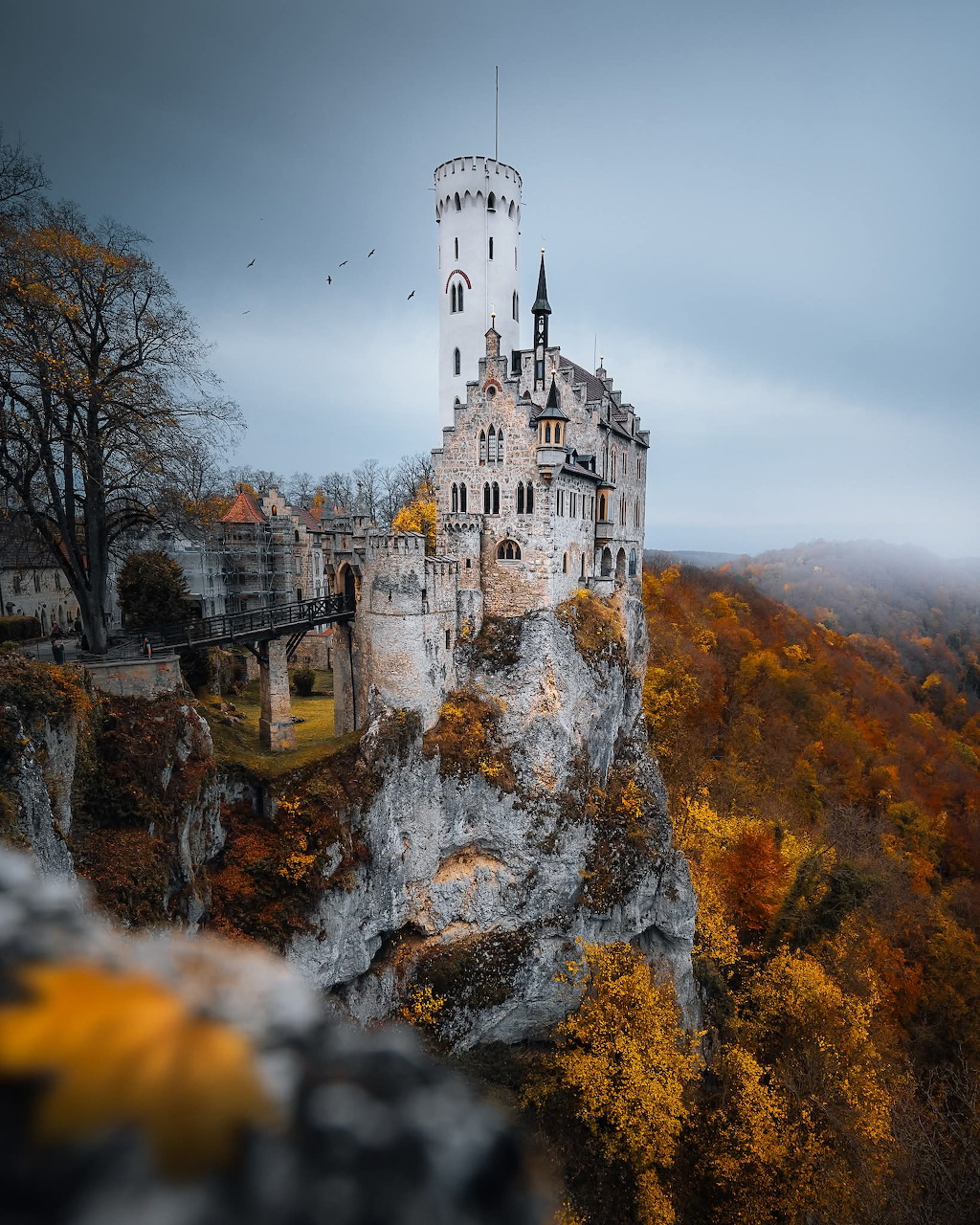 Gothic white castle atop a rocky cliff surrounded by colorful autumn trees and fog