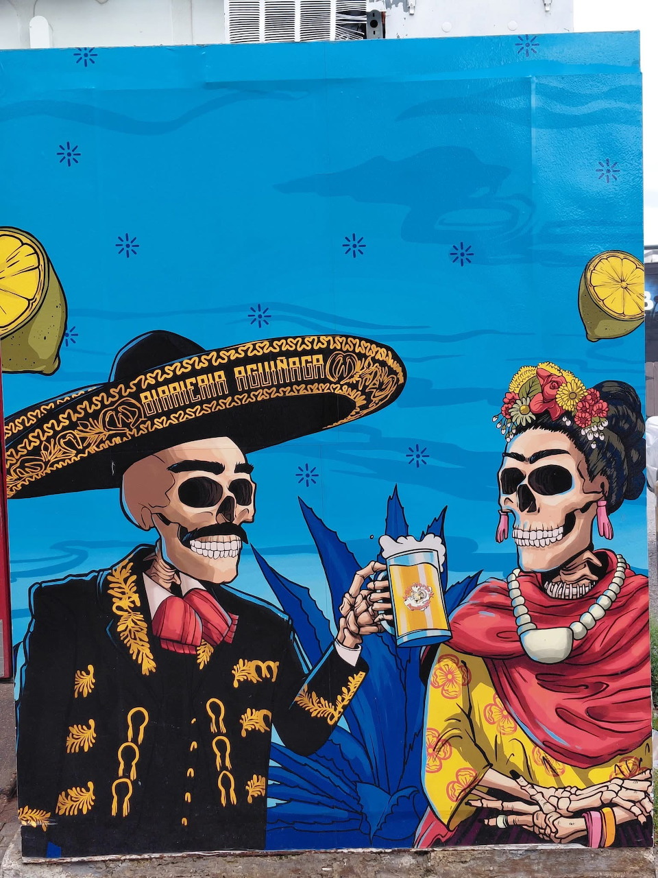 Street mural of two decorated skeletons in Mexican traditional clothing on blue wall
