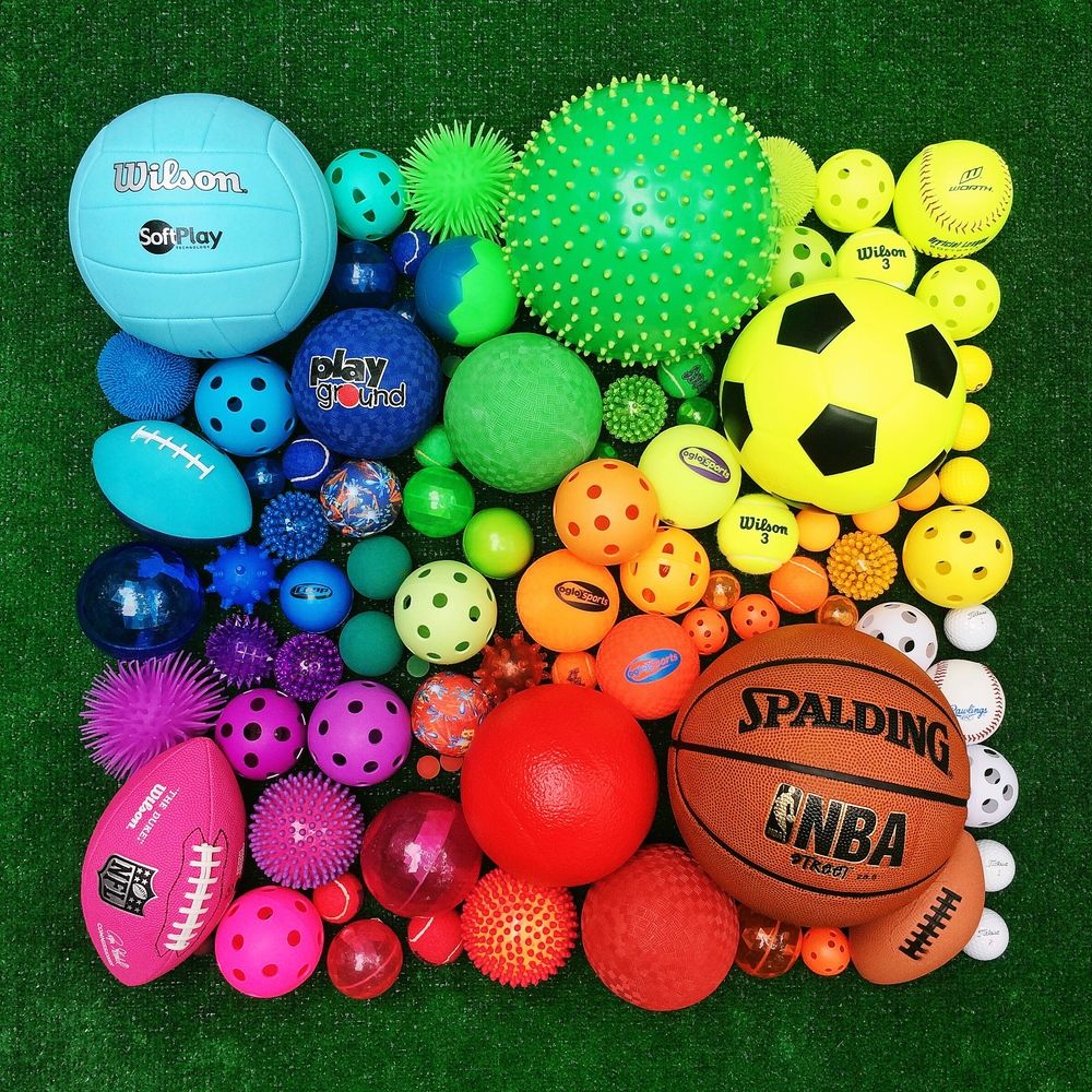 Colorful collection of sports balls and toys arranged in rainbow gradient on artificial grass