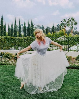Blonde woman holding out a flowing white tulle skirt while posing on a green lawn with cypress trees behind