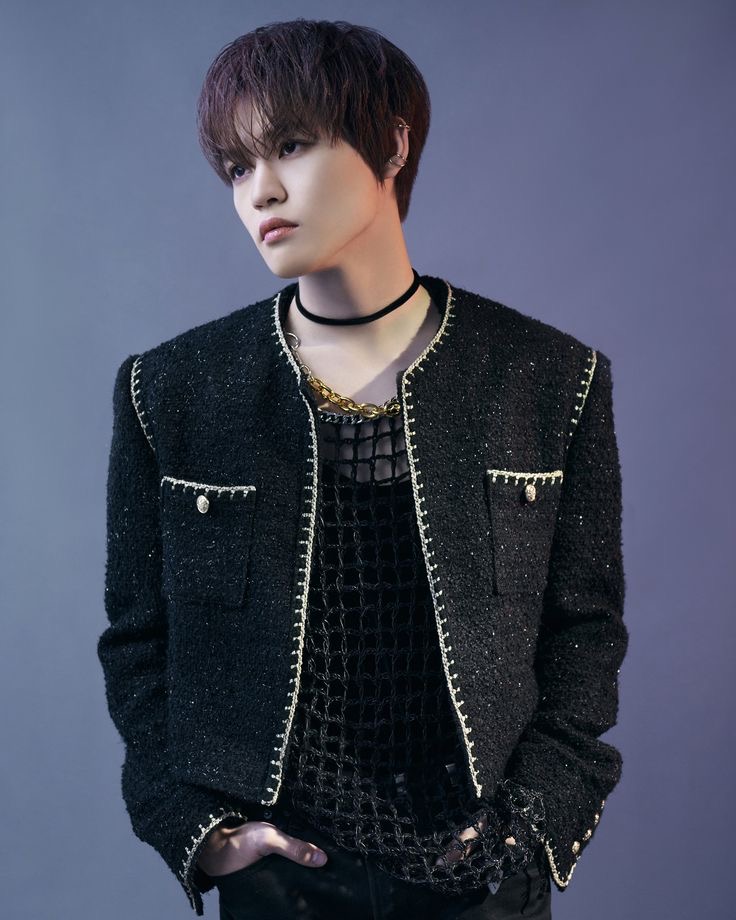 Young man with dark hair wearing a black embellished jacket with silver chains, shot against a gradient blue-gray background