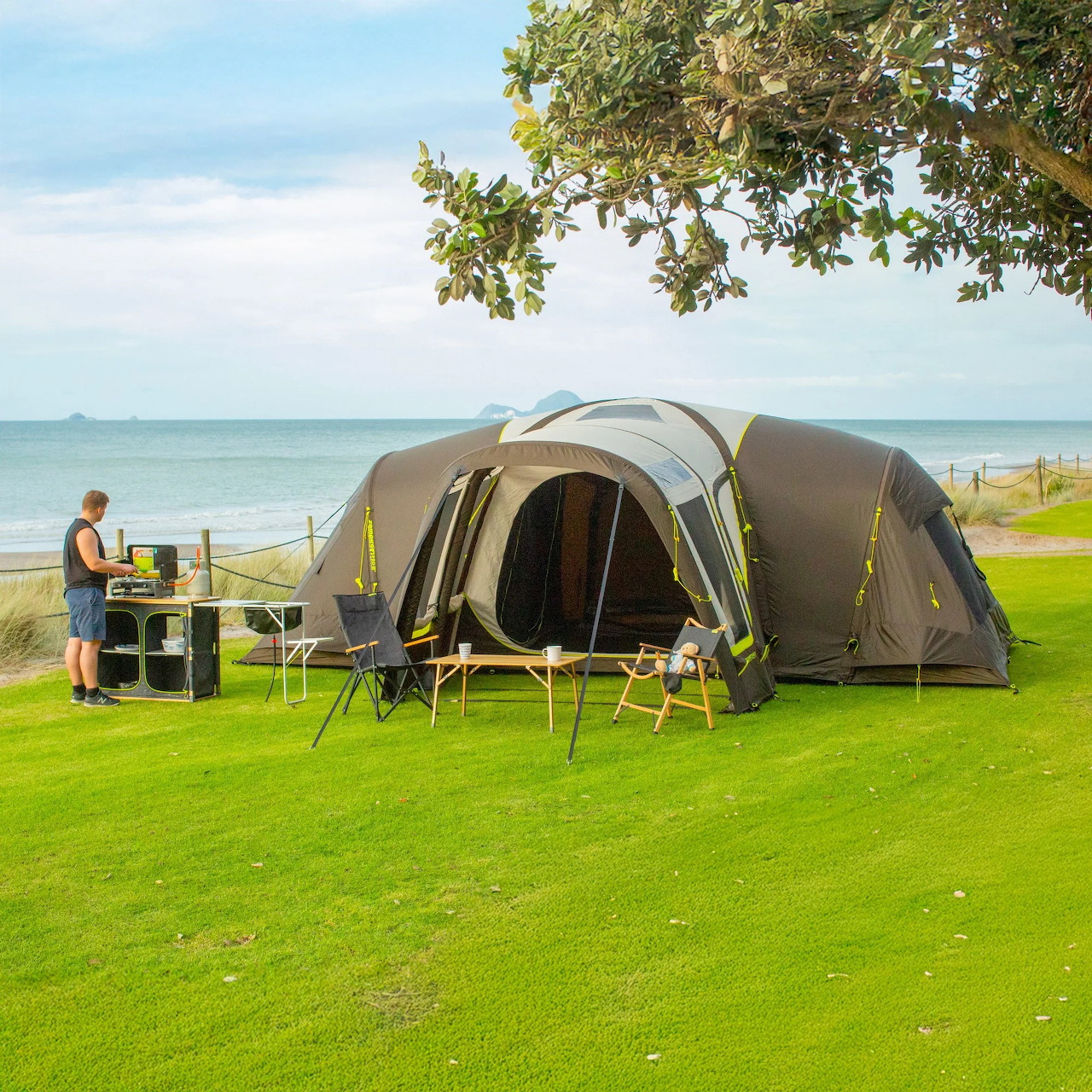 Large camping tent on a grassy coastal campsite with chairs, table, tree, and calm sea