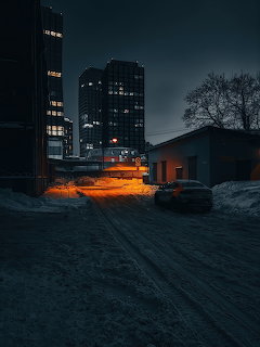 Dark snowy urban street at night with lit buildings, a parked car, and orange streetlights