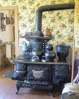 Ornate black cast iron antique wood stove with blue enamelware pots in vintage kitchen with floral wallpaper