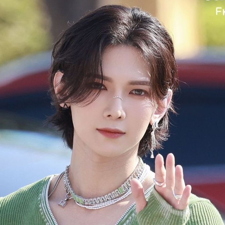 Young man with dark wavy hair in green top and silver necklace waving at camera