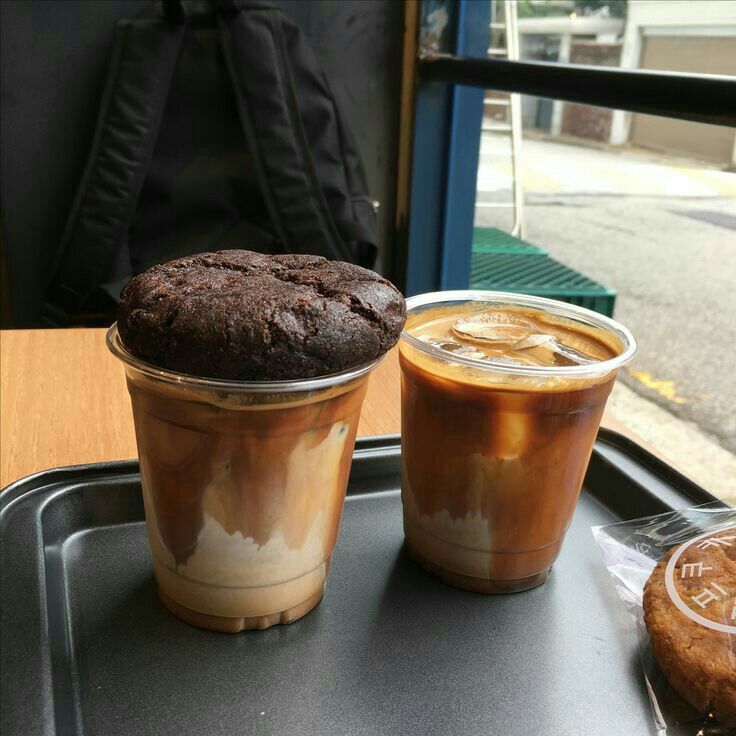 Two iced coffee drinks on a tray, one topped with a large chocolate cookie by a window