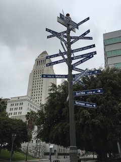 Directional signpost in downtown Los Angeles with tall buildings, palm trees, and a cloudy sky