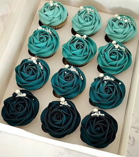 Twelve cupcakes with teal ombre rose frosting arranged in a white box, ranging from light teal to dark navy.