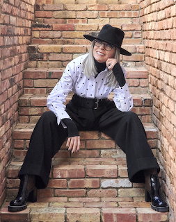 Diane Keaton sitting on brick steps in a black hat, glasses, and black-and-white outfit