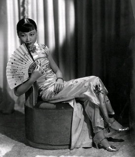Black-and-white portrait of a seated woman in a satin dress holding a fan in front of curtains.