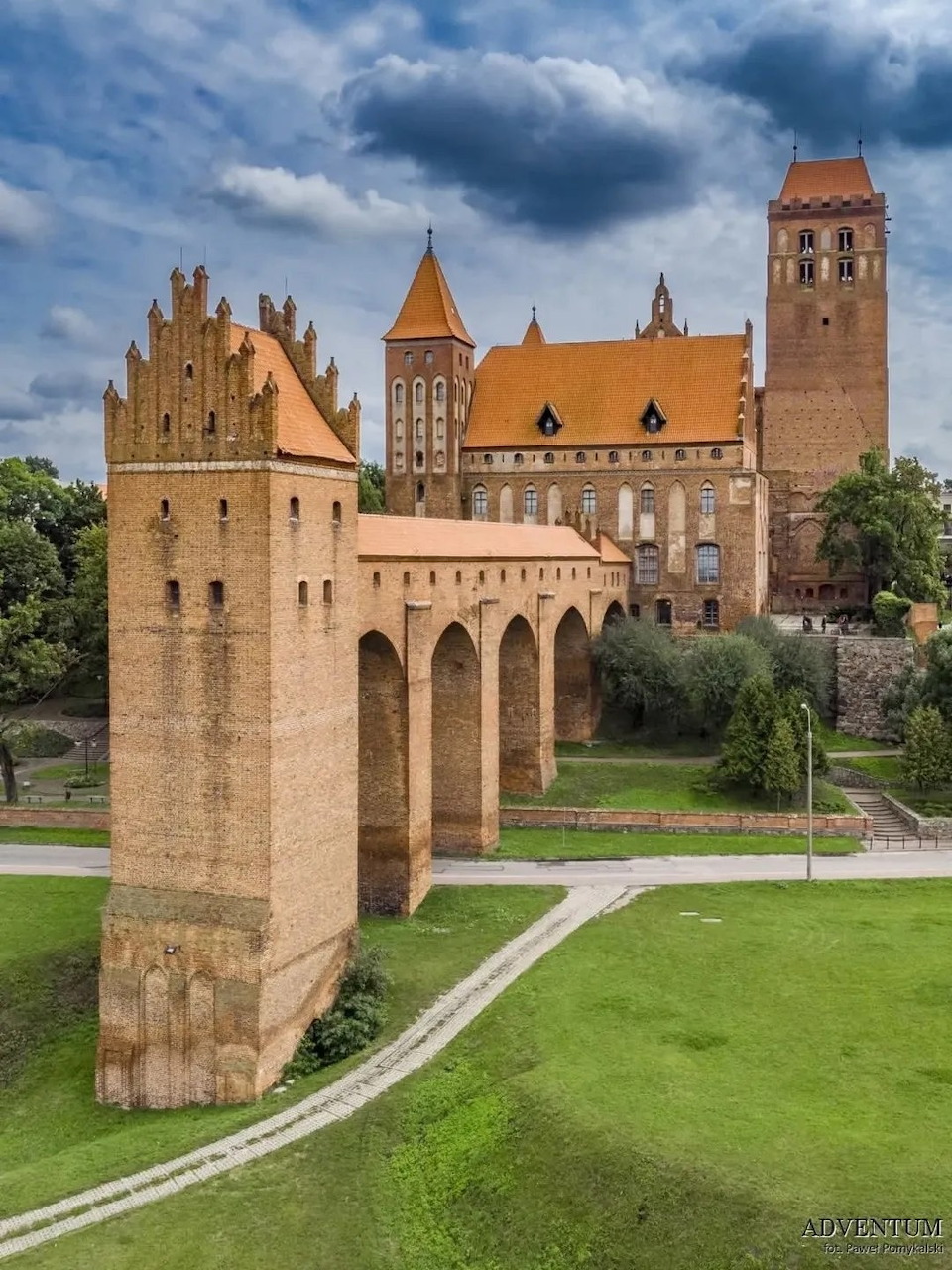 Brick castle with towers and a long elevated arcade on a grassy lawn under cloudy skies