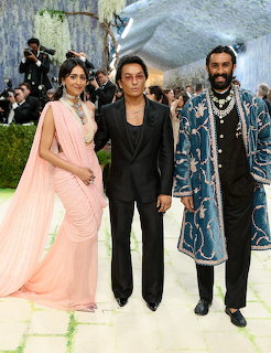 Three people posing at Met Gala: woman in pink saree, man in black suit, man in blue embroidered coat