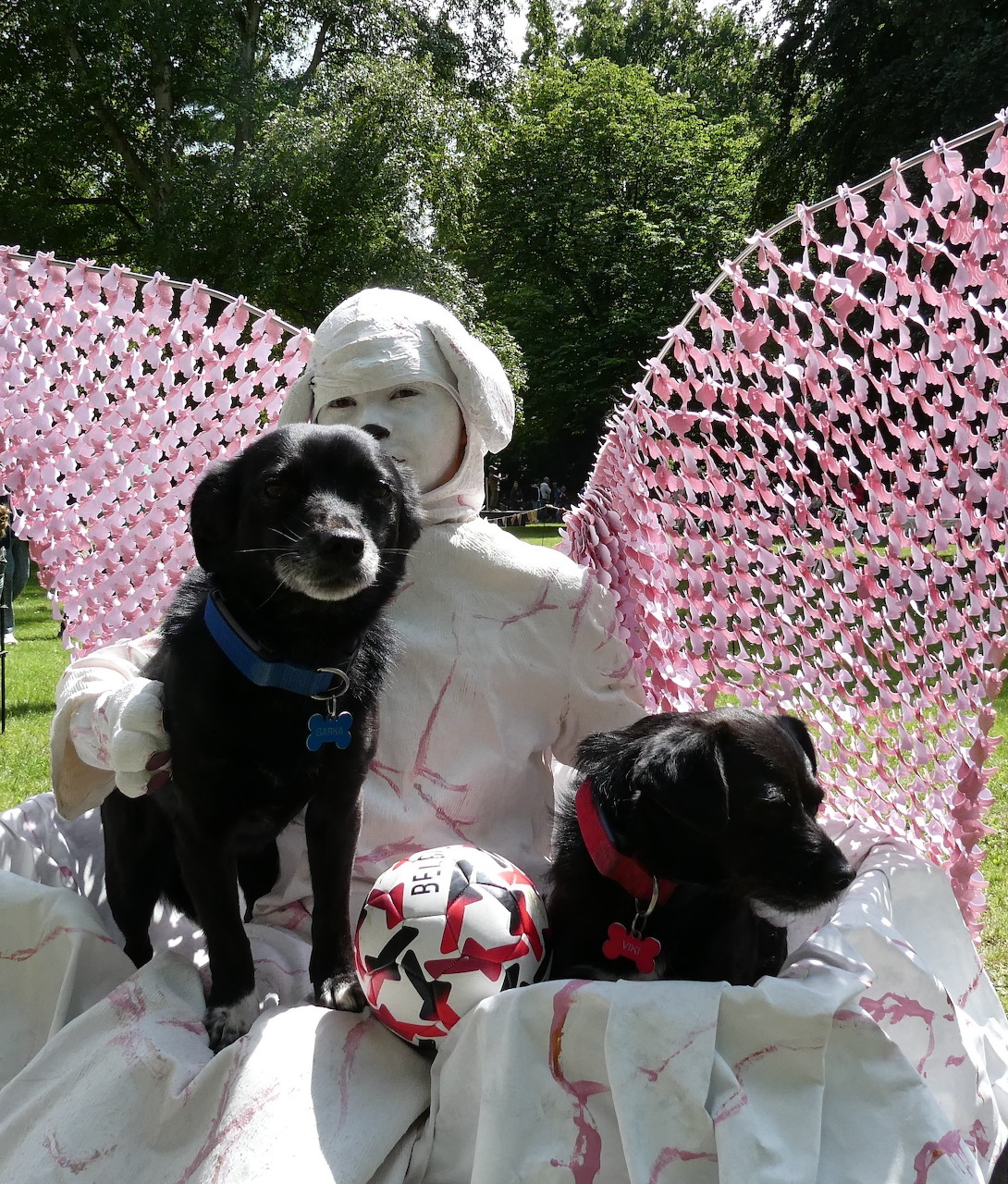 White-painted living statue performer with two black dogs and large pink wing decorations outdoors