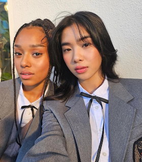 Two young women posing closely in gray blazers with white shirts and black ribbon ties.