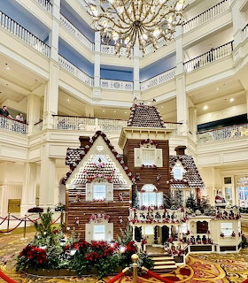 Large gingerbread house structure in ornate hotel lobby with chandelier and holiday decorations