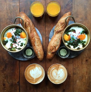 Overhead breakfast with eggs, greens, baguettes, orange juice and lattes arranged symmetrically on wood table