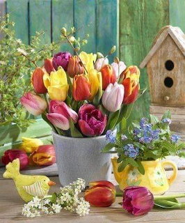 Colorful tulips in a gray pot with a yellow bird figurine, birdhouse, and small blue flowers on a wooden table