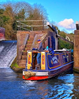Blue narrowboat passing through a canal lock with a weir and brick walls on a sunny day