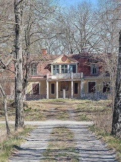 Old two-story house with balcony and red roof at the end of a dirt path lined with bare trees