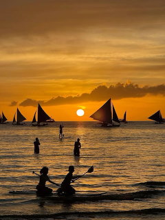 Silhouettes of sailboats and people at sunset with golden sky reflected on calm water